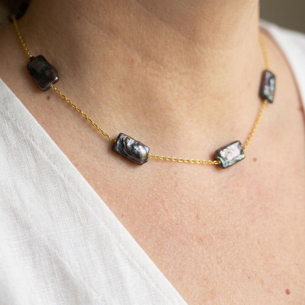 Angled close-up of dark blue rectangular baroque pearls spaced along a gold-filled chain, worn with a white top, showing their rippled surfaces and silver-green iridescence.