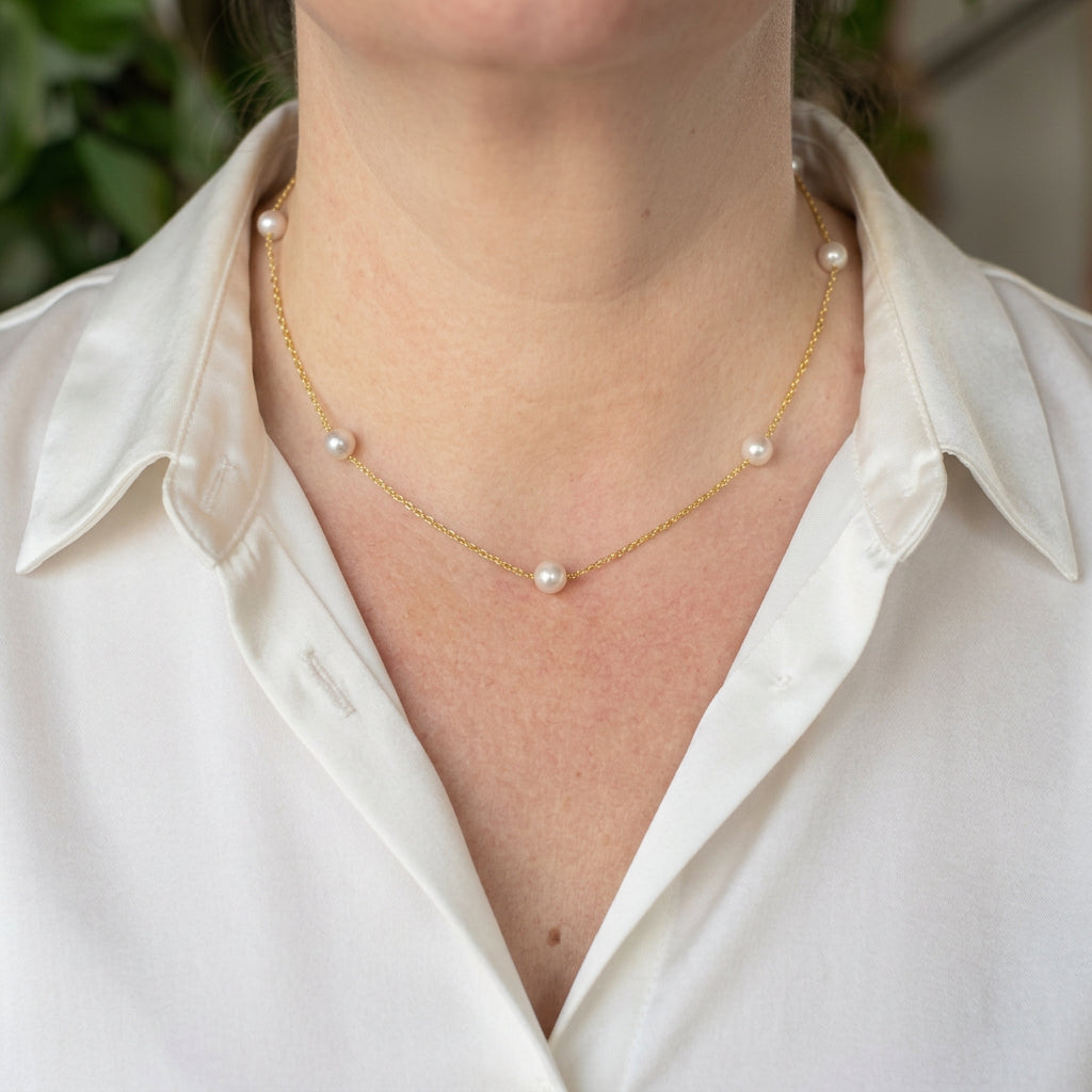 Five round white Akoya pearls spaced along a delicate gold-filled chain, worn at the collarbone with an open-neck white shirt.