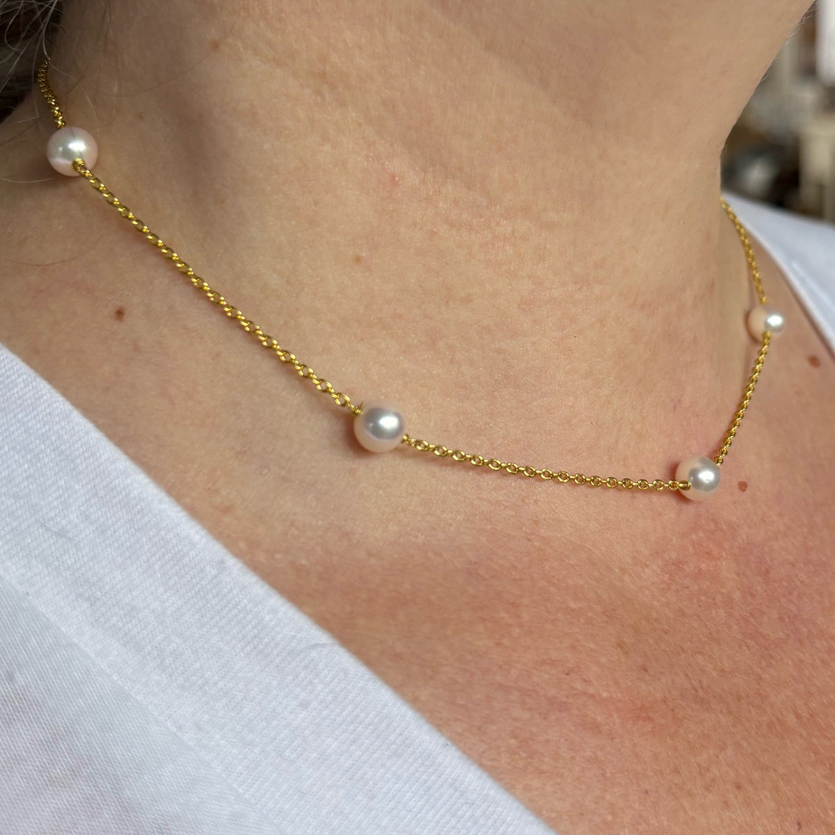 Angled close-up of an Akoya pearl station necklace worn with a white top, showing lustrous round white pearls spaced along a gold-filled chain.
