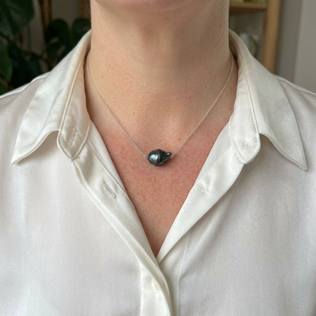 Dark blue keshi pearl set at the centre of a delicate sterling silver chain, worn with an open-neck white shirt, showing its irregular shape and deep blue-green lustre.