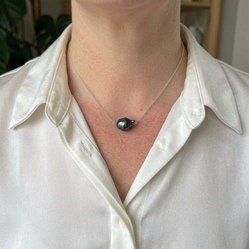 Dark blue keshi pearl set at the centre of a delicate sterling silver chain, worn with an open-neck white shirt, showing its irregular shape and deep blue-green lustre.