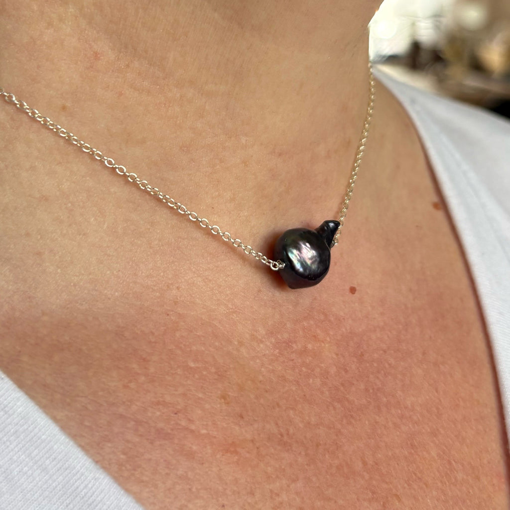 Angled close-up of a dark blue Keshi pearl necklace worn with a white top, showing the pearl’s irregular profile and reflective lustre on a fine sterling silver chain.