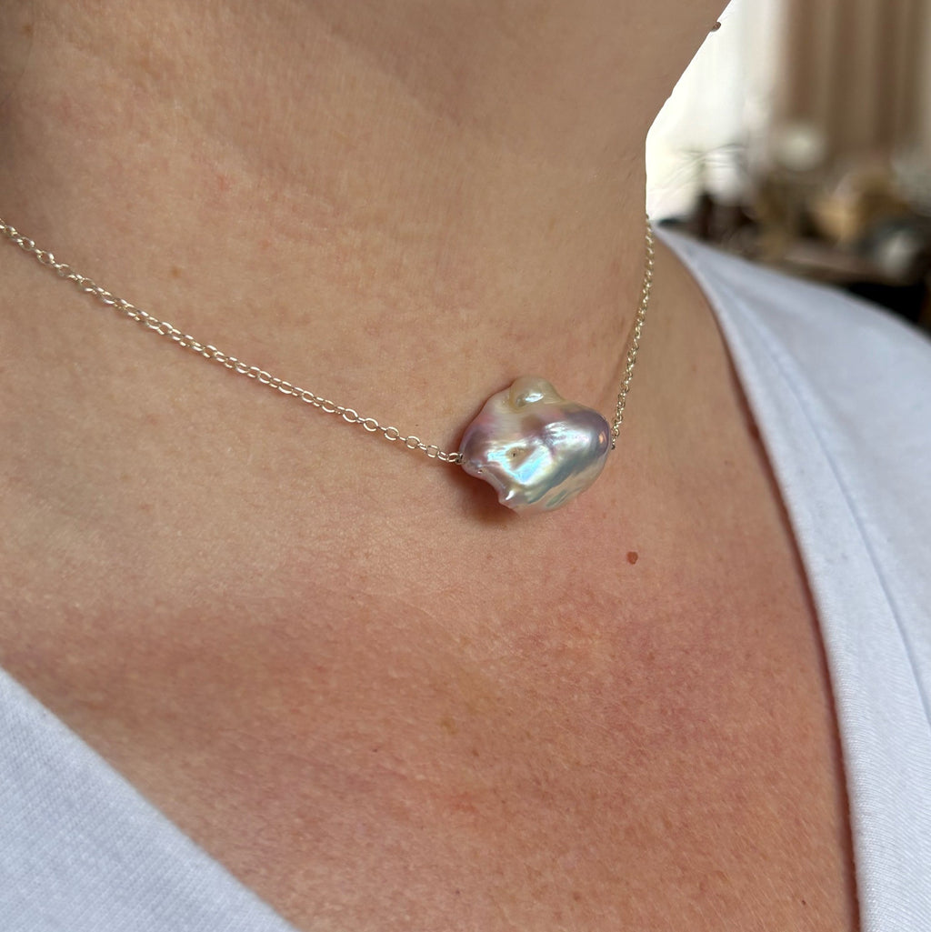 Angled close-up of a lavender Keshi pearl necklace worn with a white top, showing the pearl’s sculptural surface and blue-green iridescence on a fine sterling silver chain.⠋