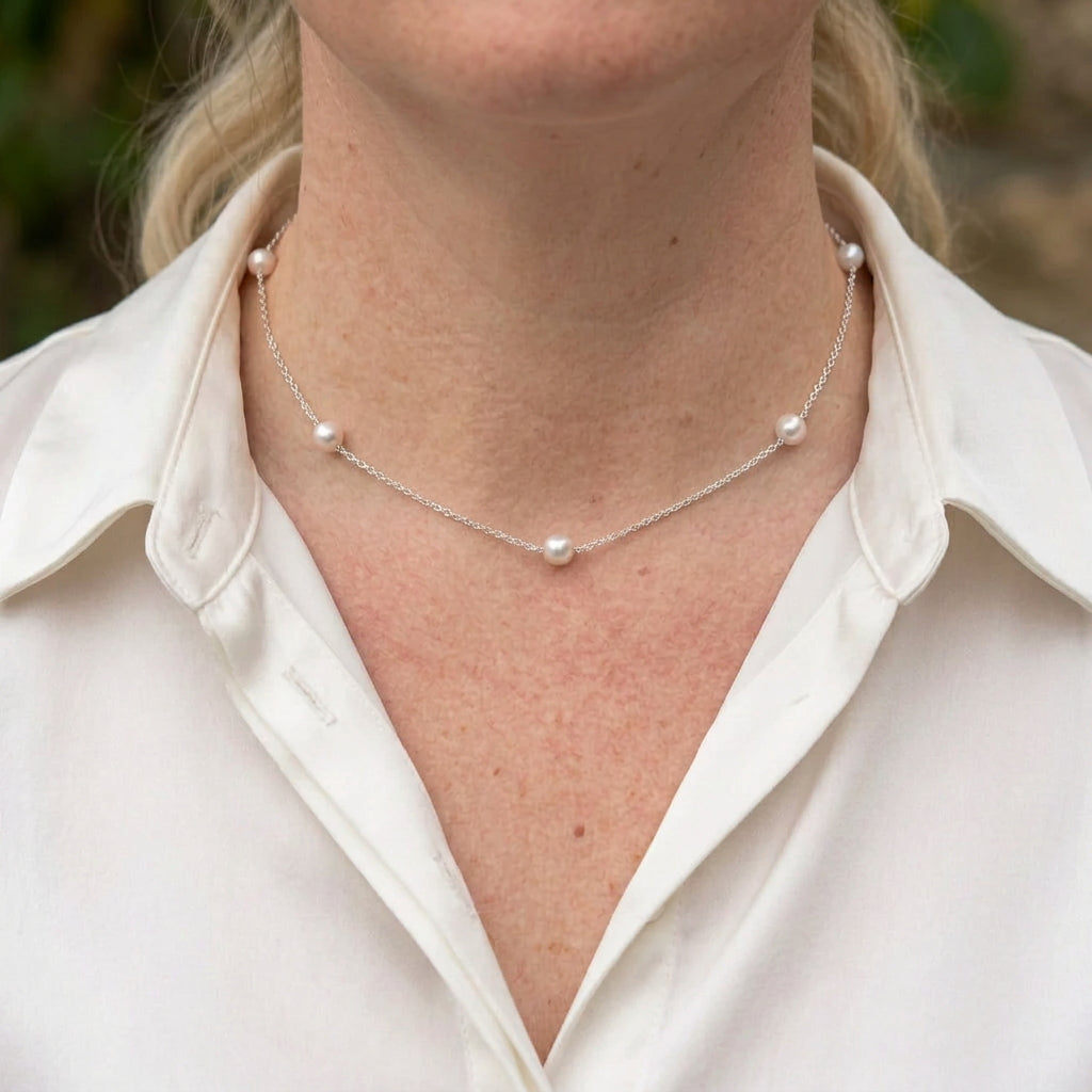 Five round white Akoya pearls spaced along a delicate sterling silver chain, worn at the collarbone with an open-neck white shirt.