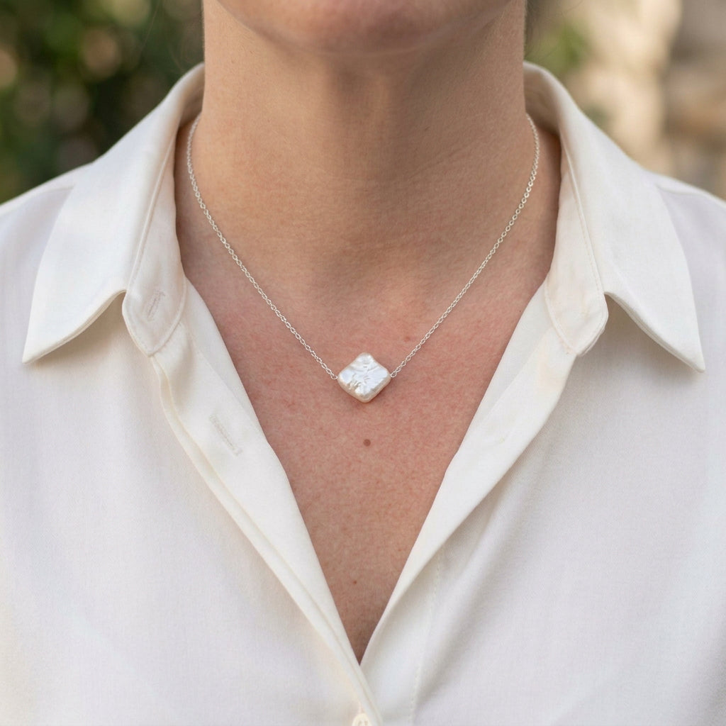 White square baroque pearl set diagonally at the centre of a delicate sterling silver chain, worn with an open-neck white shirt, showing its irregular edges and silvery iridescent surface.