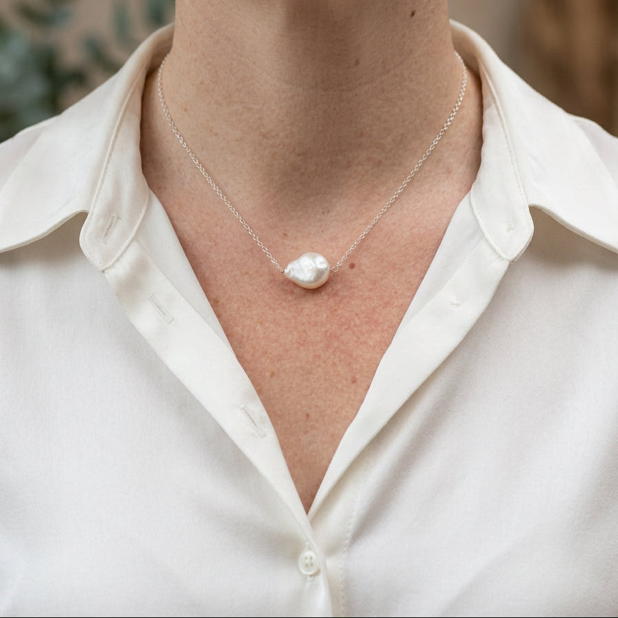 Single white baroque pearl set at the centre of a delicate sterling silver chain, worn with an open-neck white shirt, showing the pearl’s irregular shape and creamy iridescent lustre.⠋