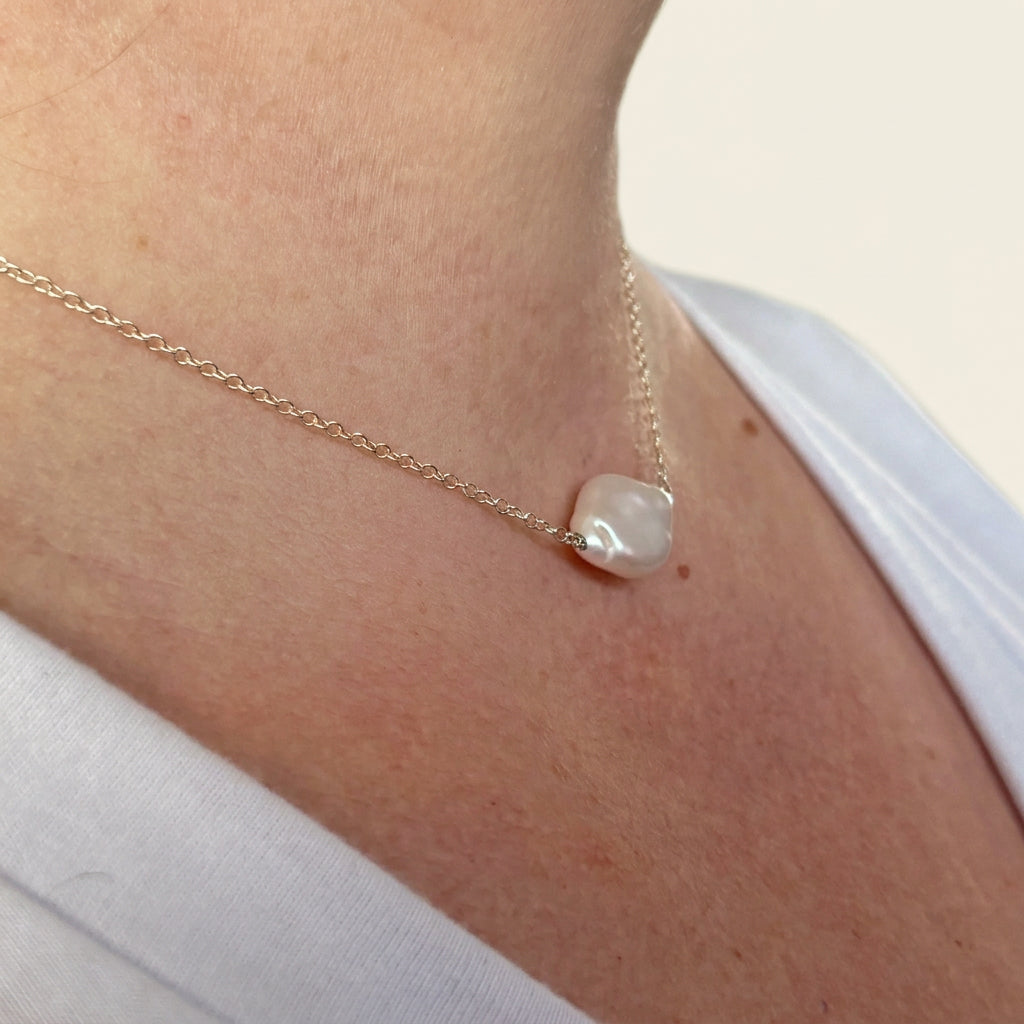 Angled close-up of a white baroque pearl necklace worn with a white top, showing the pearl’s irregular shape and silver wire-wrapped connection to a fine sterling silver chain.