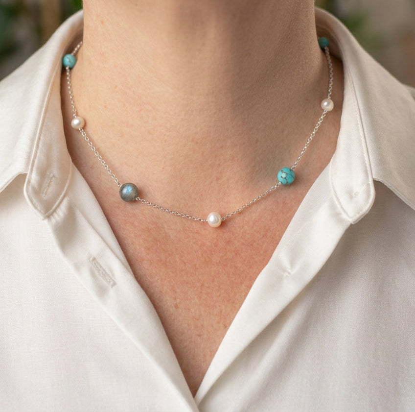 White baroque pearls alternating with irregular blue-green and grey larimar stones along a delicate chain, worn at the collarbone with an open-neck white shirt.⠋