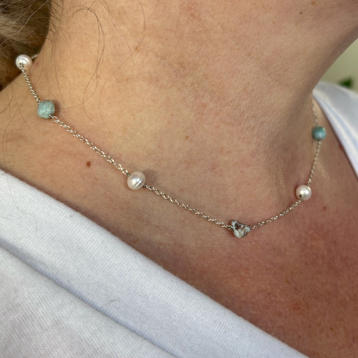 Angled close-up of a pearl and larimar station necklace worn with a white top, showing white baroque pearls alternating with pale blue, naturally patterned larimar stones on a sterling silver chain.