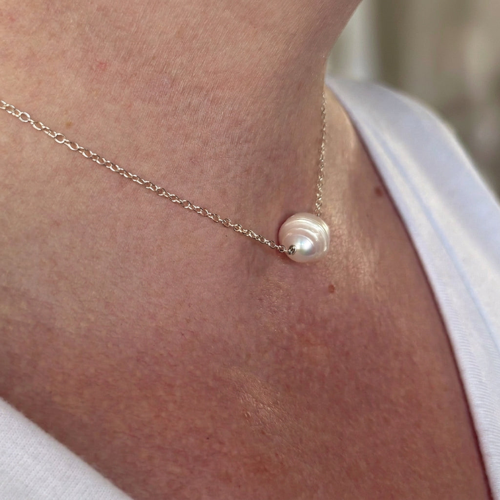 Angled close-up of a white ribbed baroque pearl necklace worn with a white top, showing the pearl’s concentric ridges and wire-wrapped connection to a fine sterling silver chain.