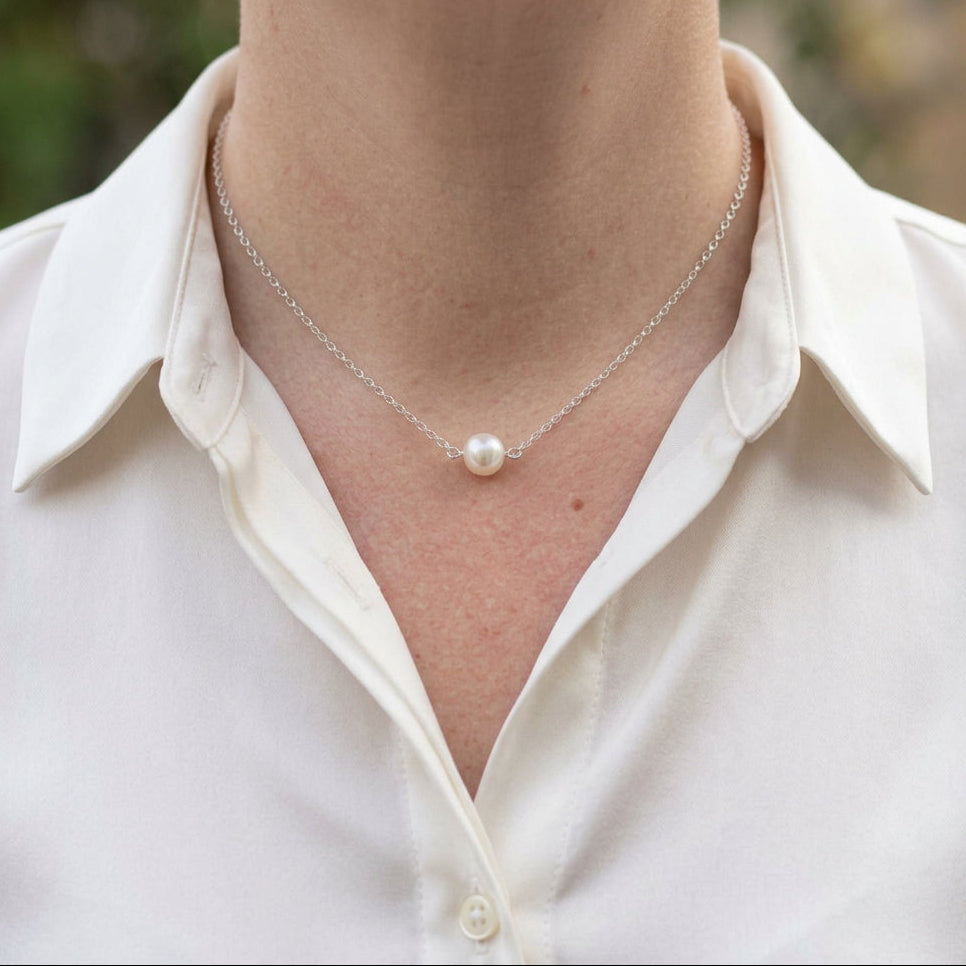 Single round white Akoya pearl set at the centre of a delicate sterling silver chain, worn at the collarbone with an open-neck white shirt.