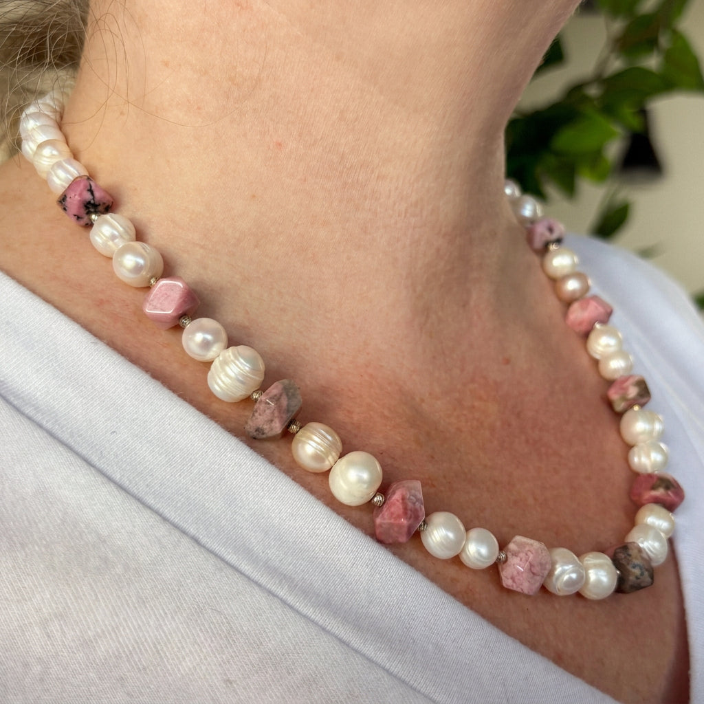 Side view of a white baroque pearl and pink opal necklace worn with a white V-neck top, showing irregular faceted pink stones with dark mottling, pairs of ridged pearls and small sterling silver spacers.