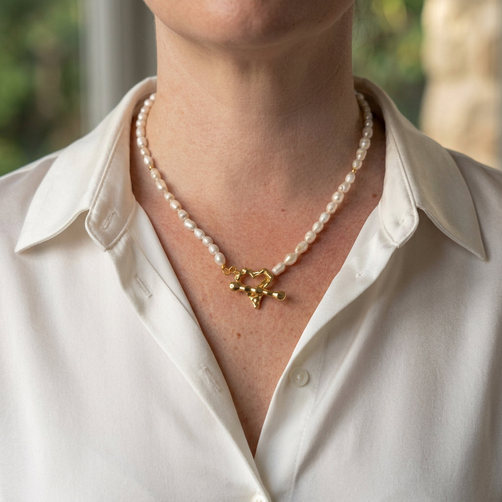 White rice pearl necklace with small gold-plated spacers and a fastened gold-plated heart-shaped front toggle clasp, worn with an open-neck white shirt.