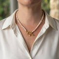 White rice pearl necklace with small gold-plated spacers and a fastened gold-plated heart-shaped front toggle clasp, worn with an open-neck white shirt.