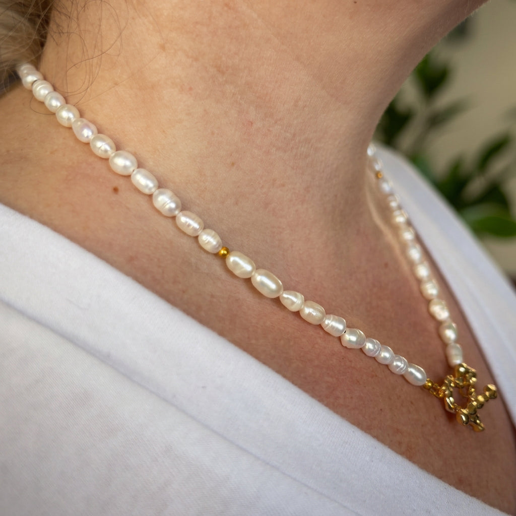Side view of a white rice-shaped baroque pearl necklace worn with a white V-neck top, showing the pearls’ irregular shapes, small gold-plated spacer beads and a front heart-shaped toggle clasp.⠋