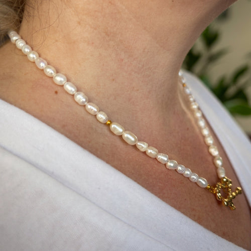 Side view of a white rice-shaped baroque pearl necklace worn with a white V-neck top, showing the pearls’ irregular shapes, small gold-plated spacer beads and a front heart-shaped toggle clasp.⠋