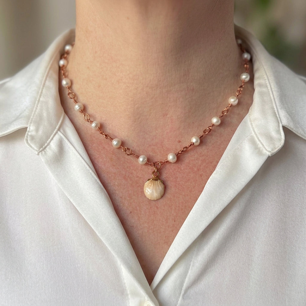White baroque pearl necklace with handmade copper wire links, worn with an open-neck white shirt, featuring a cream scallop shell pendant with a gold-coloured cap.