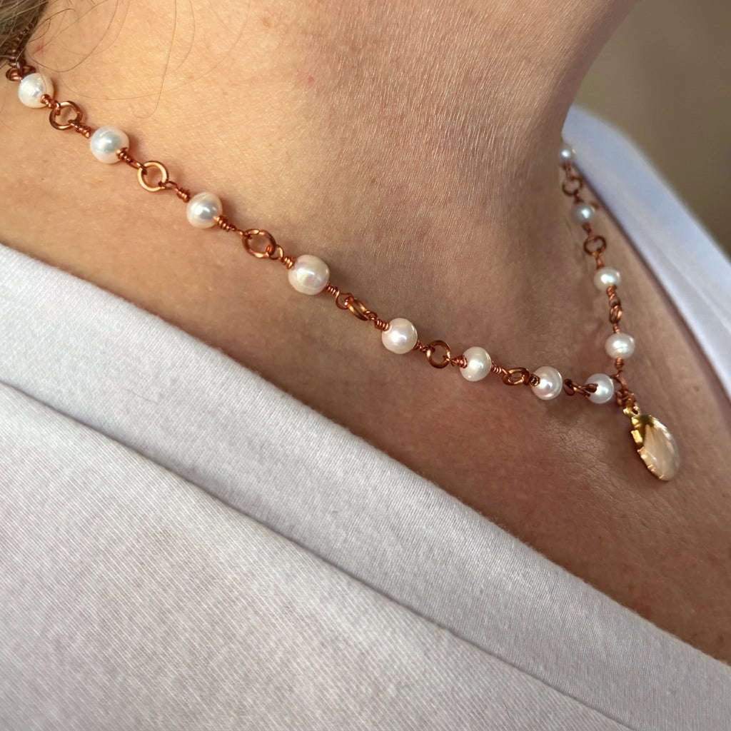Angled view of a white baroque pearl necklace worn with a white top, showing copper wire-wrapped links and a cream shell pendant hanging at the centre.