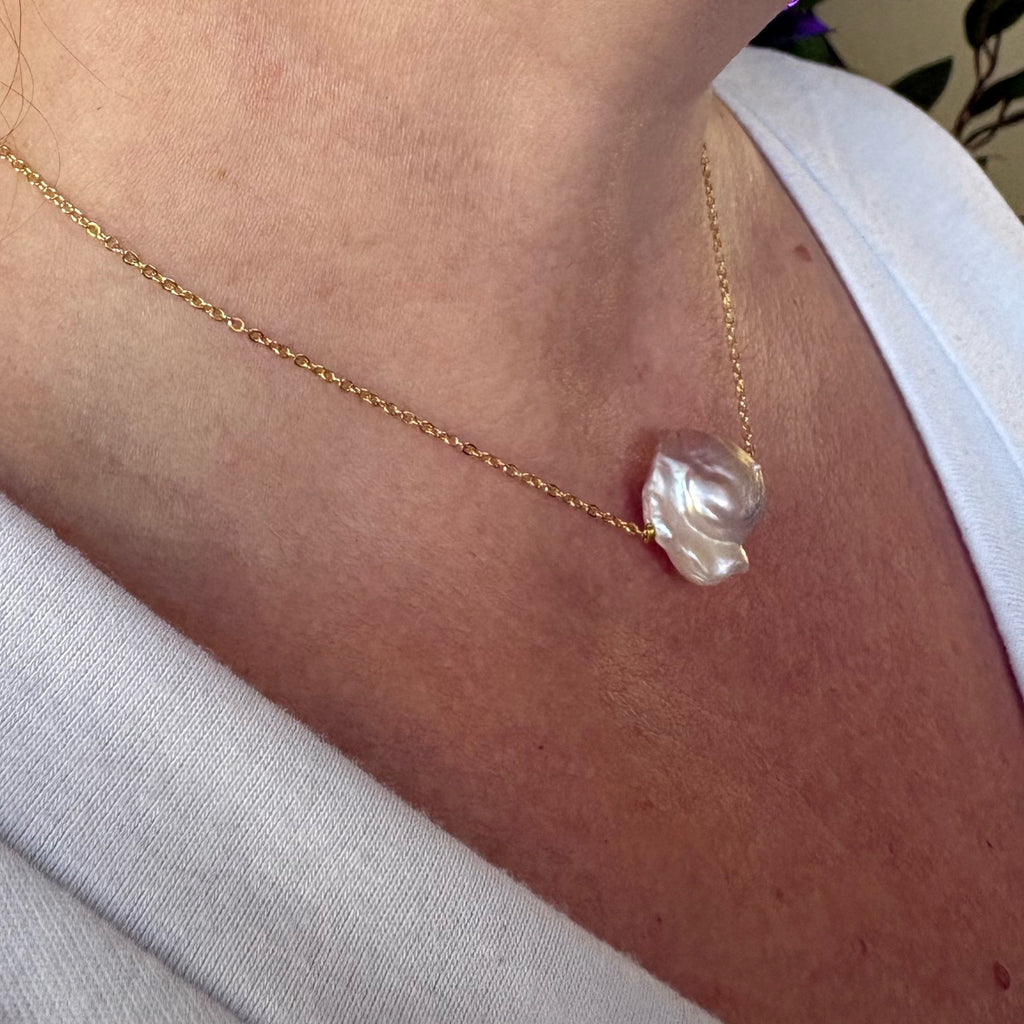 Side-angle view of a silvery-blush keshi pearl on a fine gold-over-sterling-silver chain, worn with a white V-neck top, highlighting the pearl’s sculptural folds and bright nacre lustre.