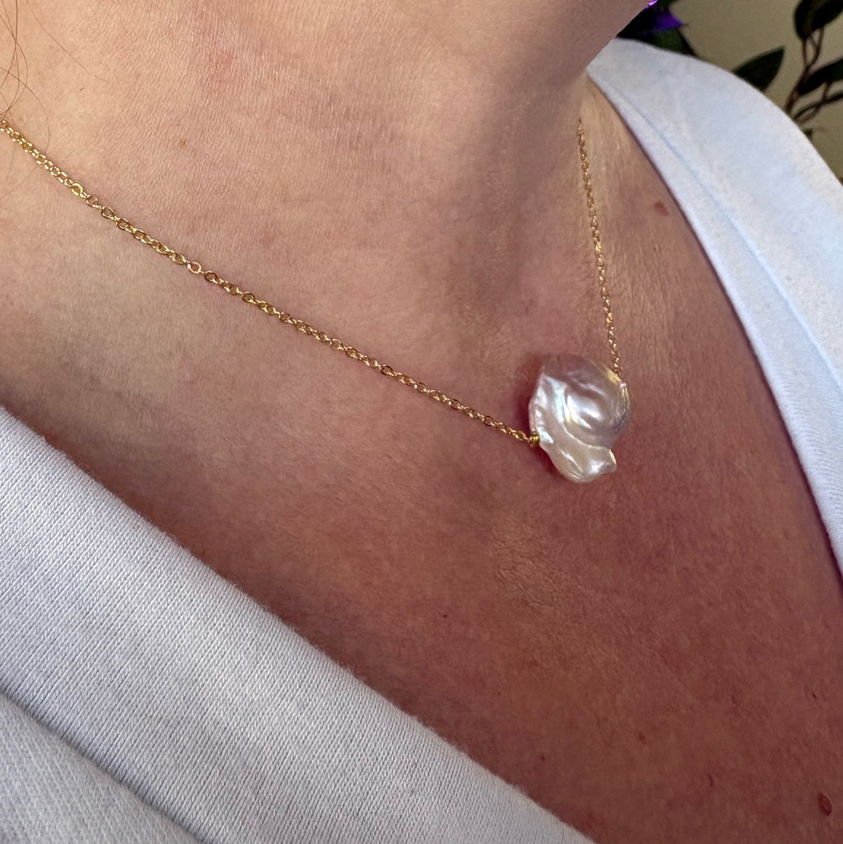 Side-angle view of a silvery-blush keshi pearl on a fine gold-over-sterling-silver chain, worn with a white V-neck top, highlighting the pearl’s sculptural folds and bright nacre lustre.