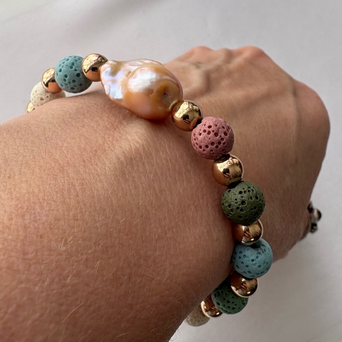 Angled close-up of a bracelet worn on a wrist, showing a large peach-gold keshi pearl with porous dusty pink, green and teal lava beads alternating with polished gold-plated spacers.