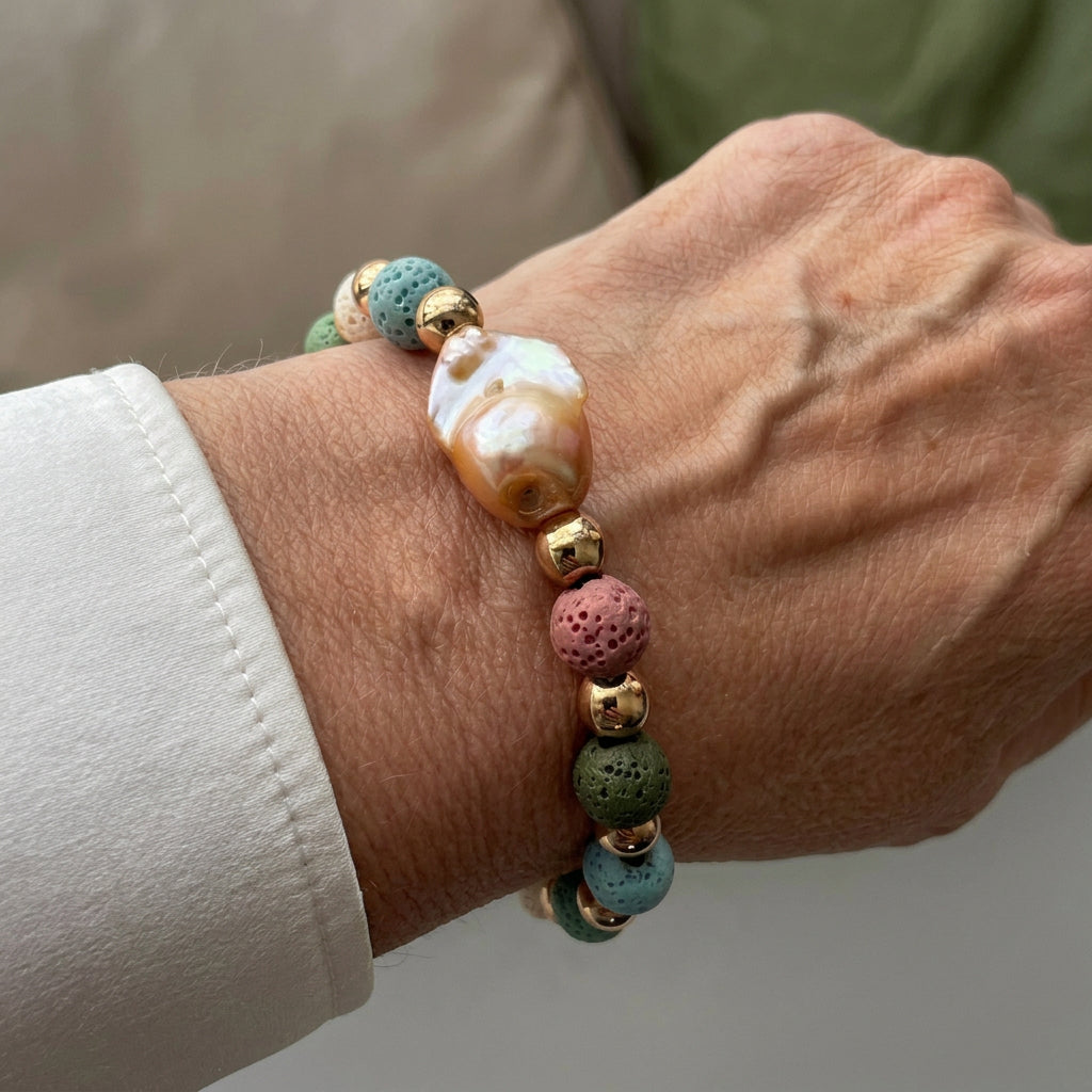 Bracelet with a large peach-gold keshi pearl, porous pink, blue and green lava beads and gold-coloured spacers, worn on the wrist beside a white silk shirt cuff.⠋