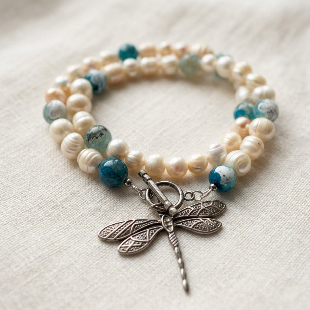 White baroque pearl necklace coiled on cream linen, with blue glass accent beads, a fastened silver-coloured toggle clasp and a large stainless steel dragonfly pendant with patterned wings.