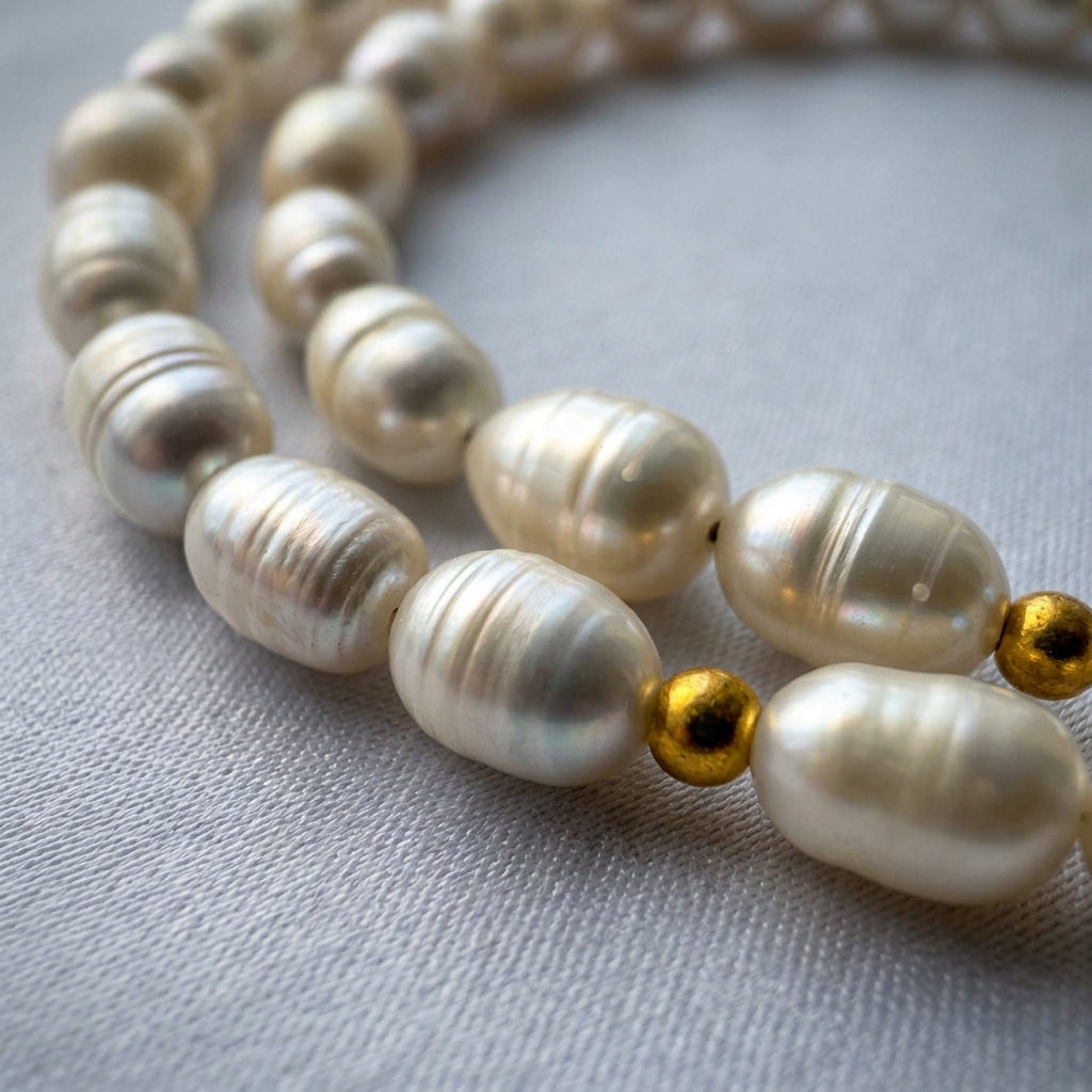 Close-up of white rice-shaped baroque pearls and a small gold-plated spacer bead on grey linen, showing the pearls’ irregular shapes, natural ridges and soft iridescent lustre.