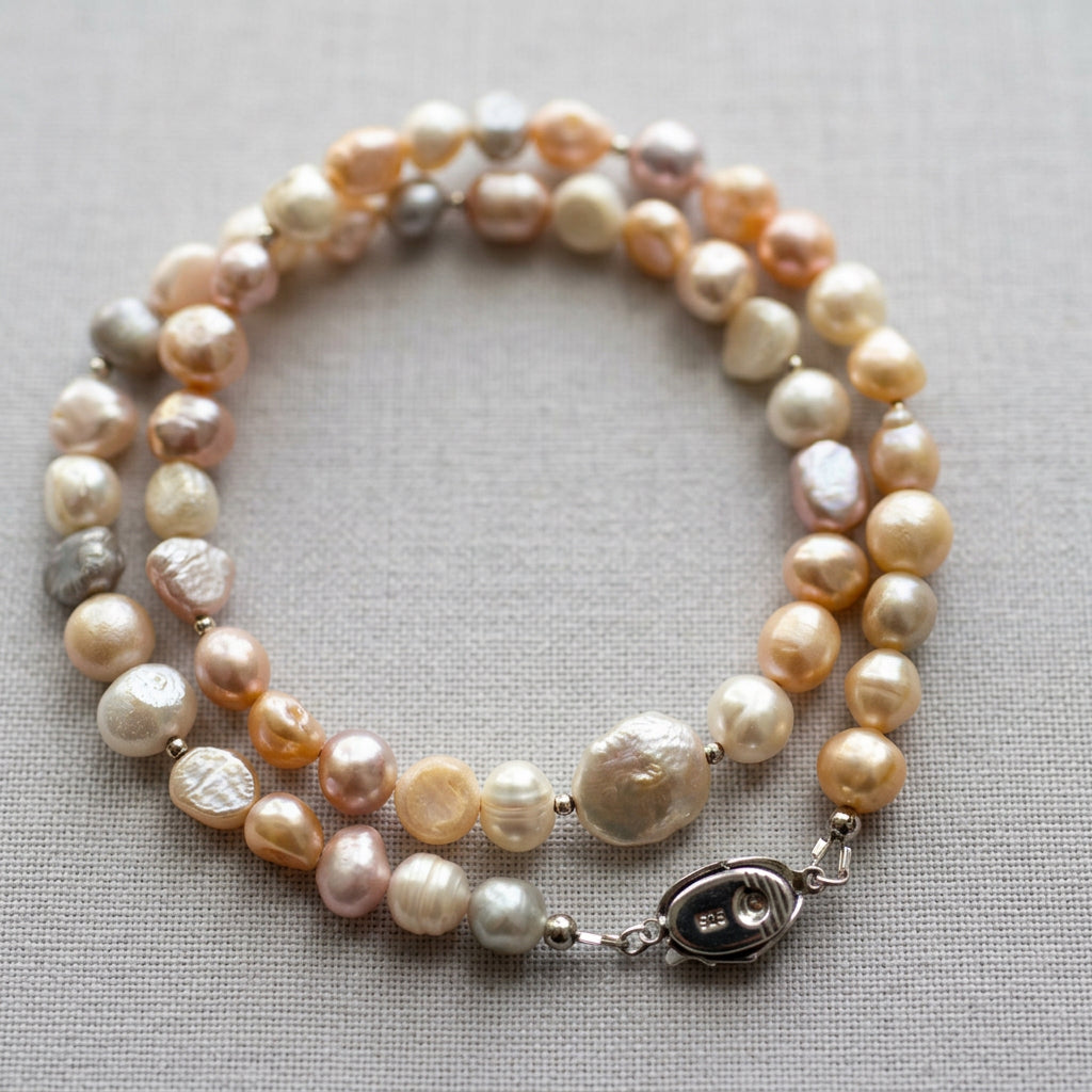 Multicoloured baroque pearl necklace loosely coiled on grey linen, showing cream, peach, pale gold and lavender pearls in varied rounded and flattened shapes, with sterling silver spacers and a fastened clasp stamped 925.