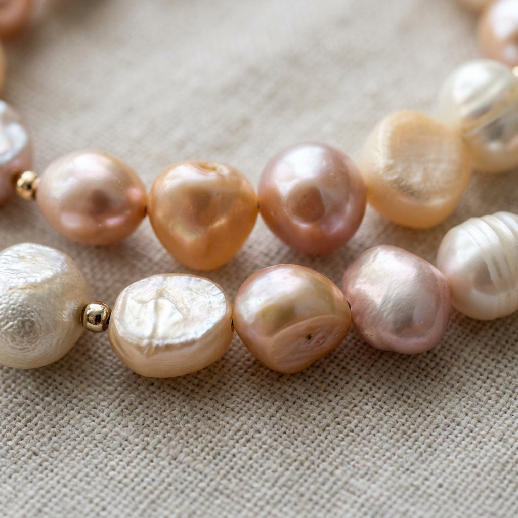 Close-up of peach, pale gold, lavender and creamy white baroque pearls on grey linen, showing rounded and flattened shapes, natural surface textures and small sterling silver spacers.