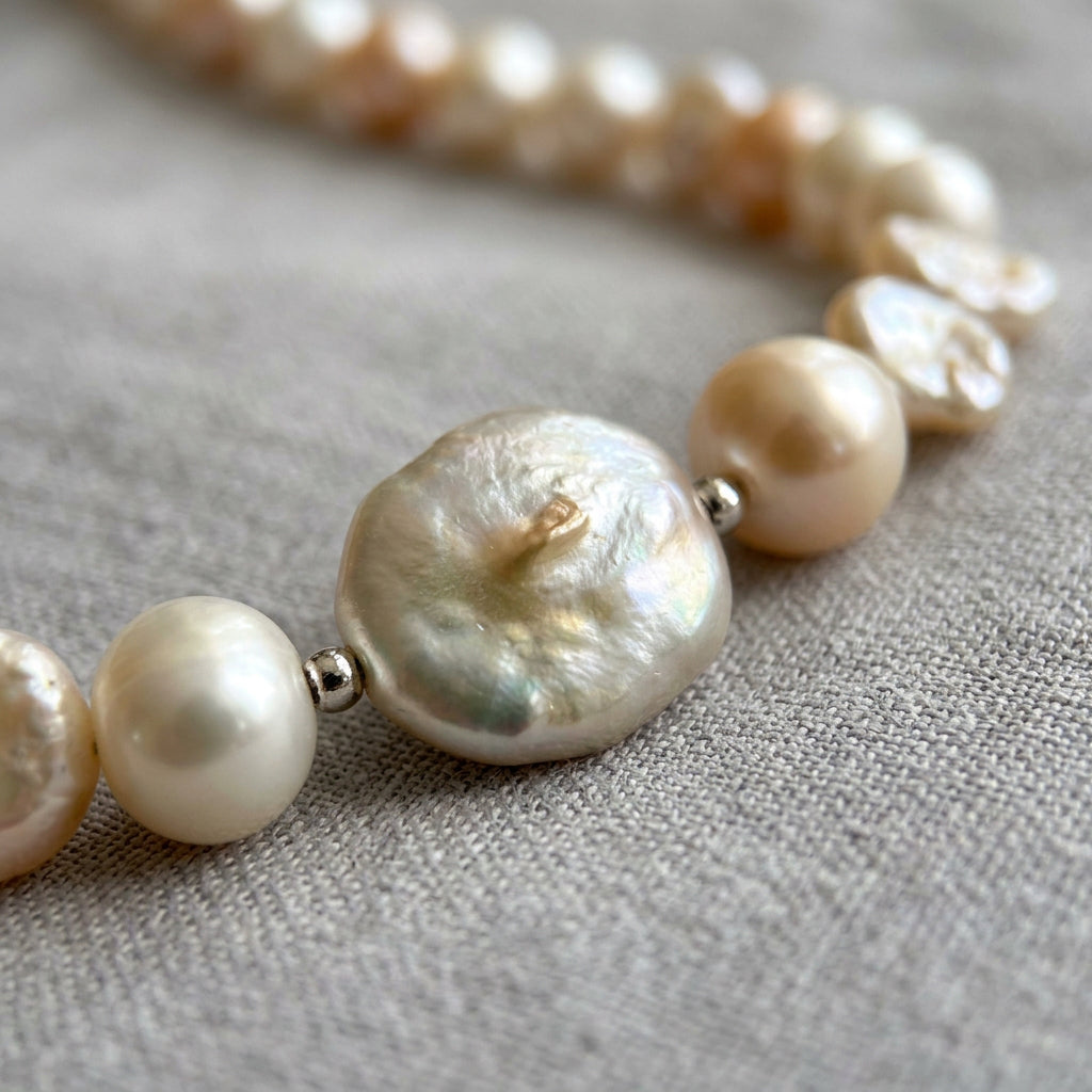 Close-up of a large flattened cream baroque pearl with a textured, iridescent surface, flanked by sterling silver spacers and smaller cream and peach pearls on grey linen.