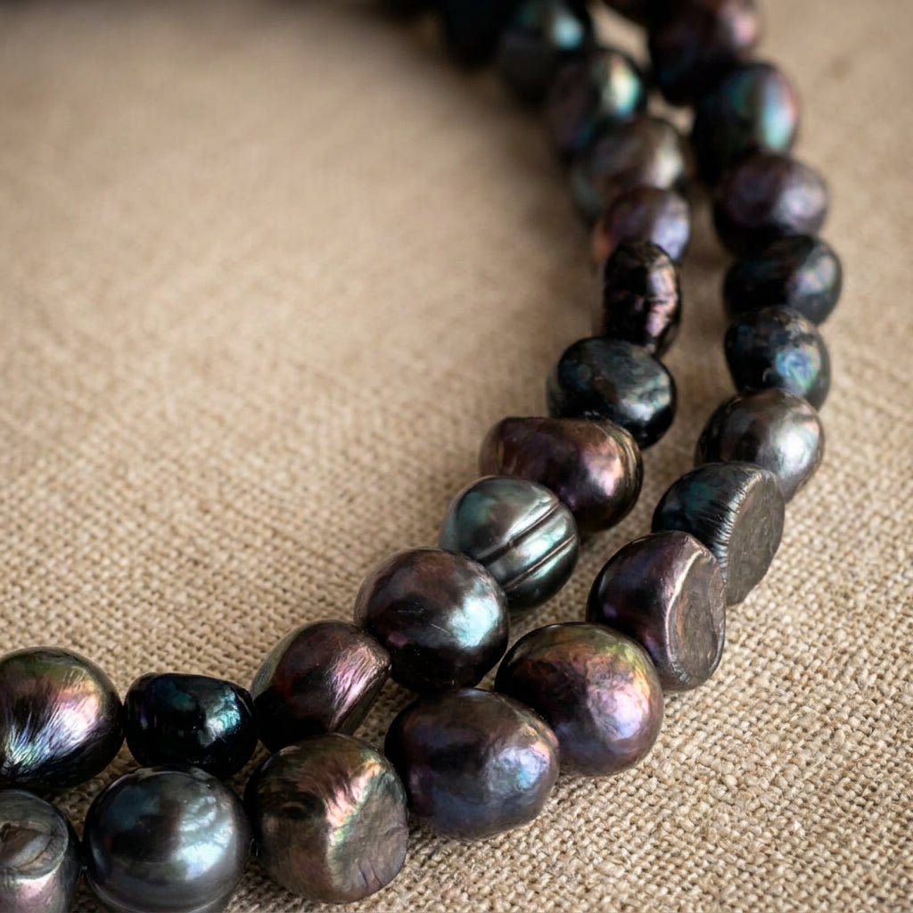 Close-up of loosely coiled dark baroque pearls on cream linen, showing irregular rounded and flattened shapes, natural surface ridges and blue, teal, purple and bronze reflections.⠋