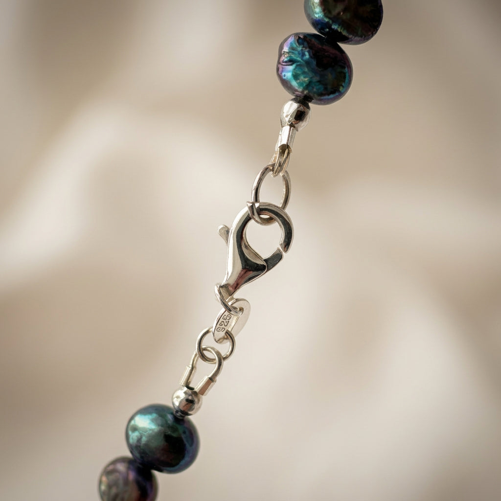 Close-up of a fastened sterling silver lobster clasp and a small tag stamped 925, joining dark baroque pearls with teal-blue reflections against a softly blurred cream background.