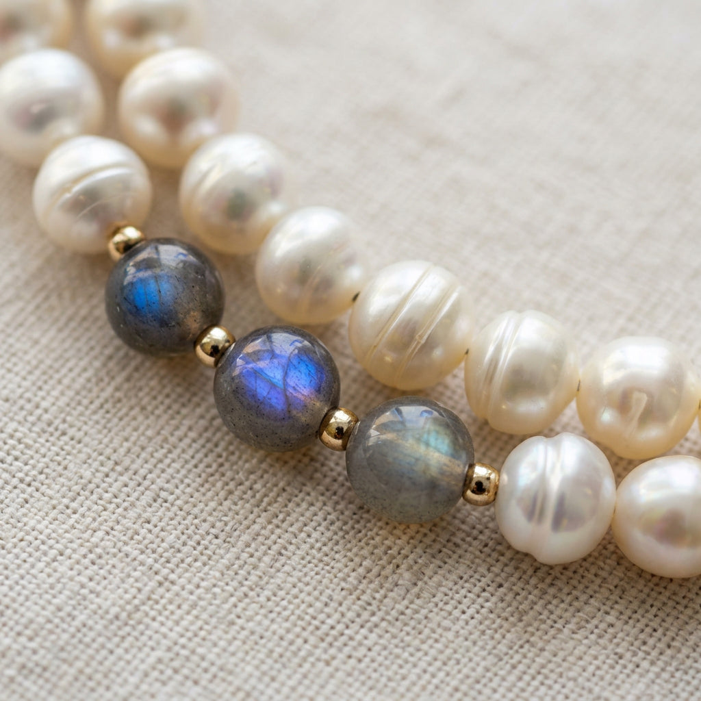 Close-up of three faceted grey labradorite beads with vivid blue flashes, separated by small sterling silver spacers beside lustrous white baroque pearls on cream linen.
