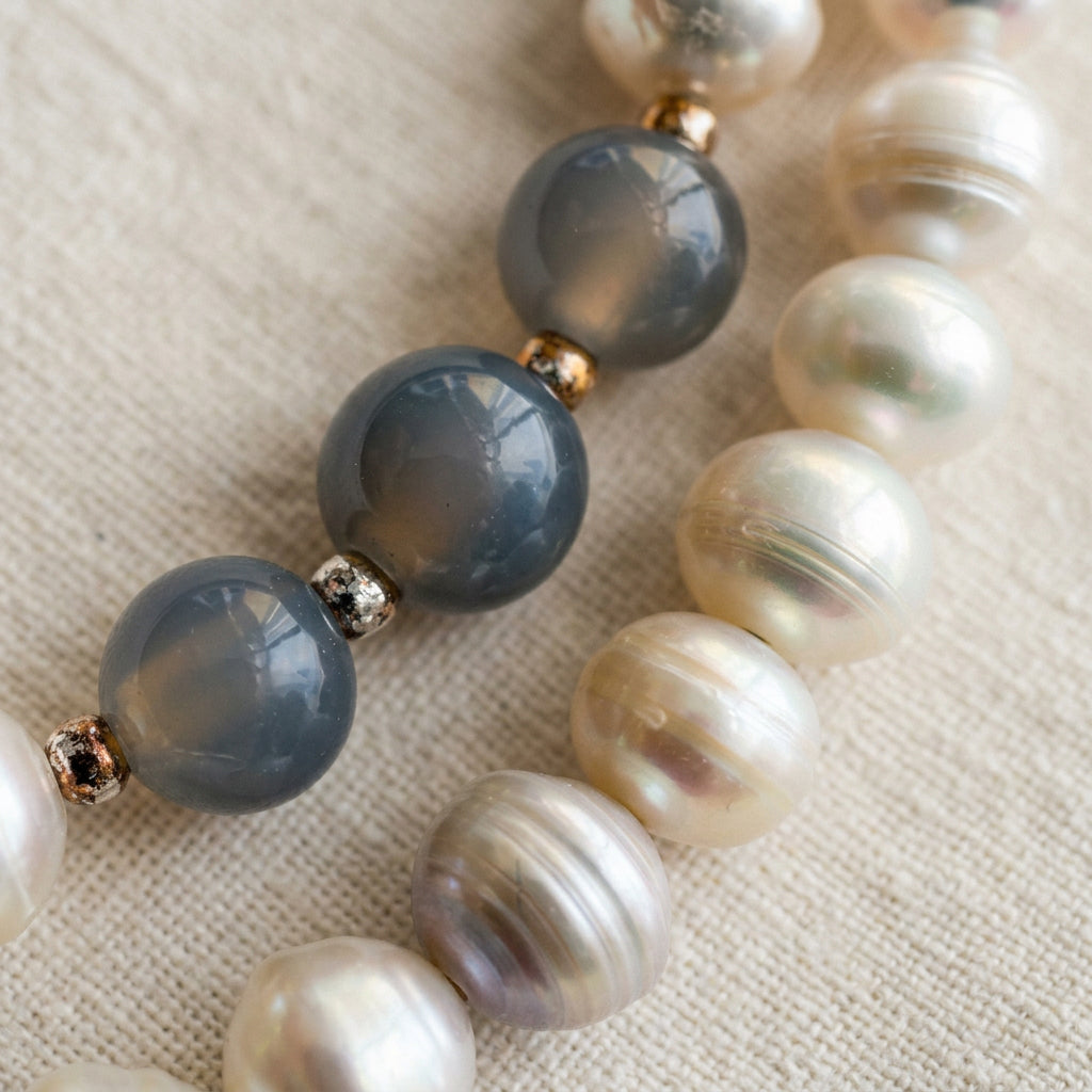 Close-up of three faceted grey labradorite beads separated by textured sterling silver spacers, beside creamy white baroque pearls with natural ridges and soft iridescence on cream linen.