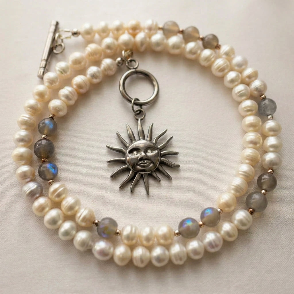 White baroque pearl and grey labradorite necklace coiled on cream fabric, showing blue flashes in the stones, a stainless steel sun face pendant and an unfastened toggle clasp with the ring and bar separated.