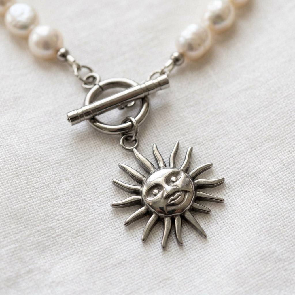 Close-up of a stainless steel sun face pendant with sculpted features and wavy rays, hanging beneath a fastened toggle clasp between white baroque pearls on cream linen.