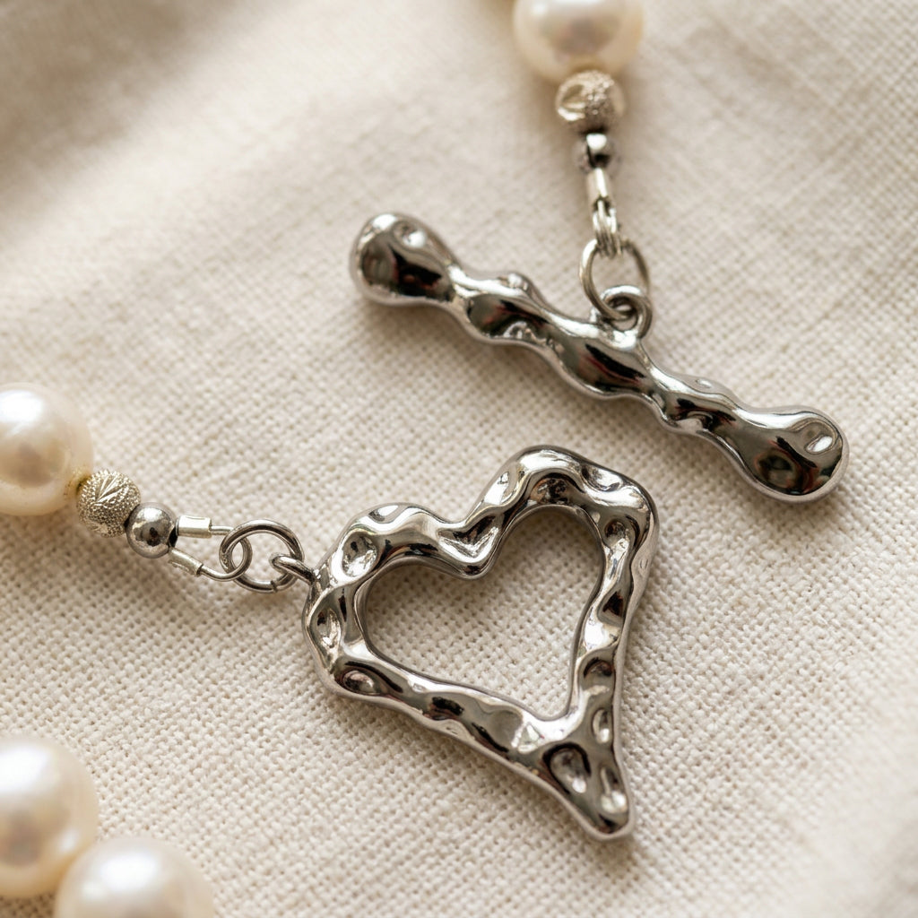 Close-up of an unfastened stainless steel heart-shaped toggle clasp with a rippled surface, showing the separate heart ring and bar, sterling silver accent beads and creamy white pearls on cream linen.