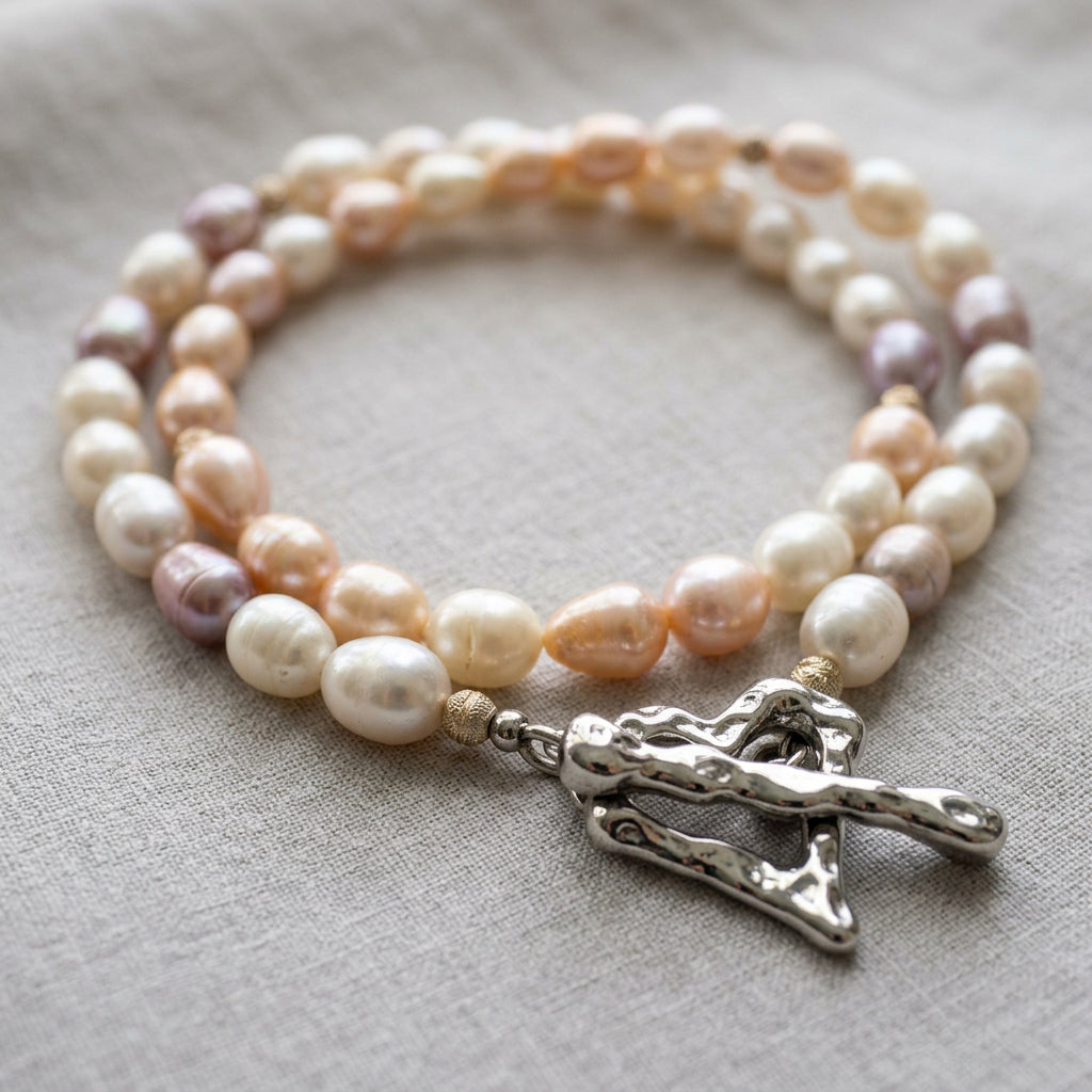 Creamy white, peach-pink and mauve rice pearl necklace loosely coiled on grey linen, with sterling silver accent beads and a fastened, rippled stainless steel heart-shaped toggle clasp in the foreground.