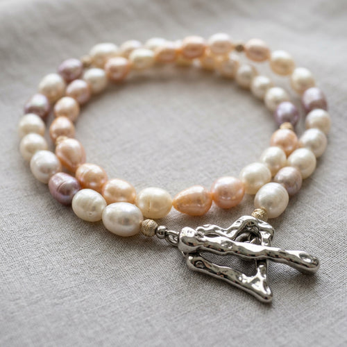 Creamy white, peach-pink and mauve rice pearl necklace loosely coiled on grey linen, with sterling silver accent beads and a fastened, rippled stainless steel heart-shaped toggle clasp in the foreground.
