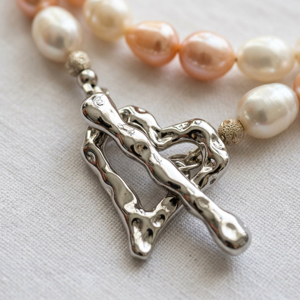 Close-up of a fastened stainless steel heart-shaped toggle clasp with a rippled, polished surface, beside sterling silver accent beads and creamy white and peach-pink baroque pearls on cream fabric.
