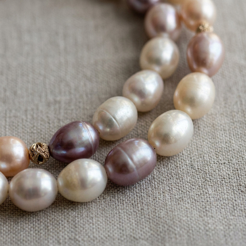 Close-up of creamy white, mauve and peach-pink rice-shaped baroque pearls with a textured sterling silver accent bead on grey linen, showing natural surface ridges and iridescent lustre.