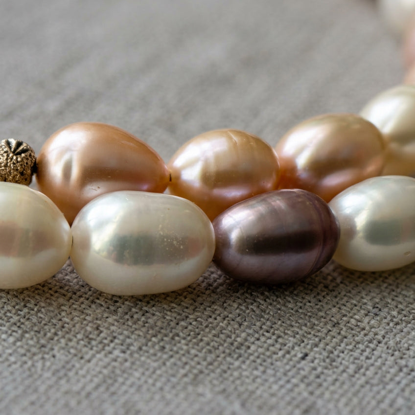 Close-up of creamy white, peach-pink and mauve rice-shaped baroque pearls on grey linen, showing their natural ridges, irregular shapes and soft iridescent lustre.