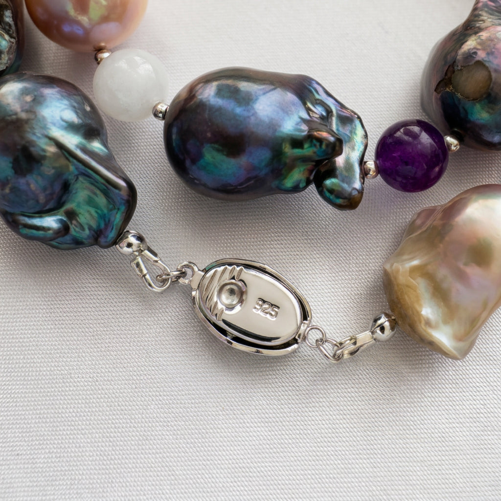 Close-up of an oval sterling silver clasp stamped 925, connecting dark iridescent and golden-cream keshi pearls, with a purple gemstone bead nearby on cream fabric.