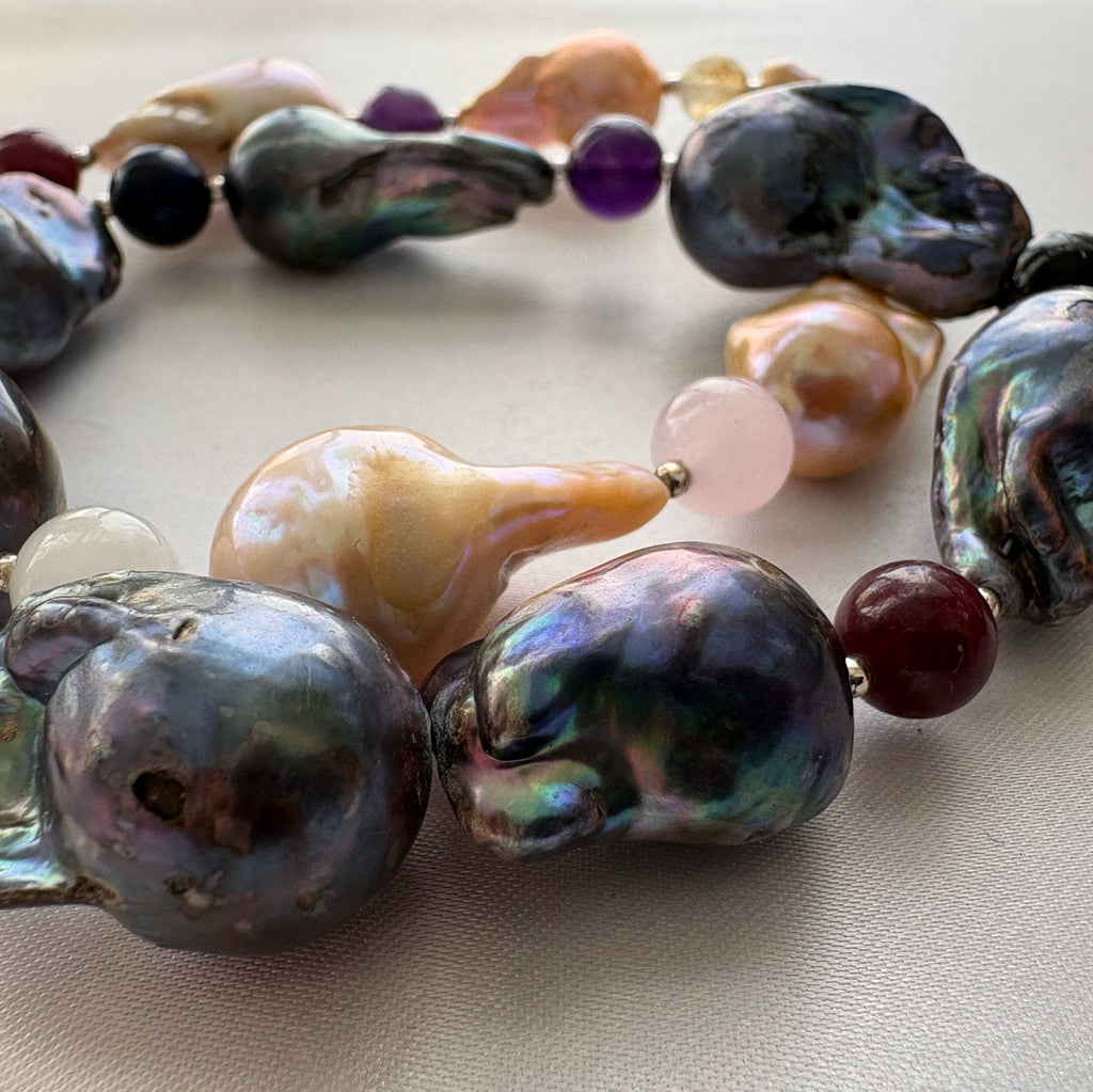 Close-up of dark blue-grey keshi pearls with green and purple iridescence, golden-cream pearls and small multicoloured gemstone beads, arranged in overlapping curves on cream fabric.
