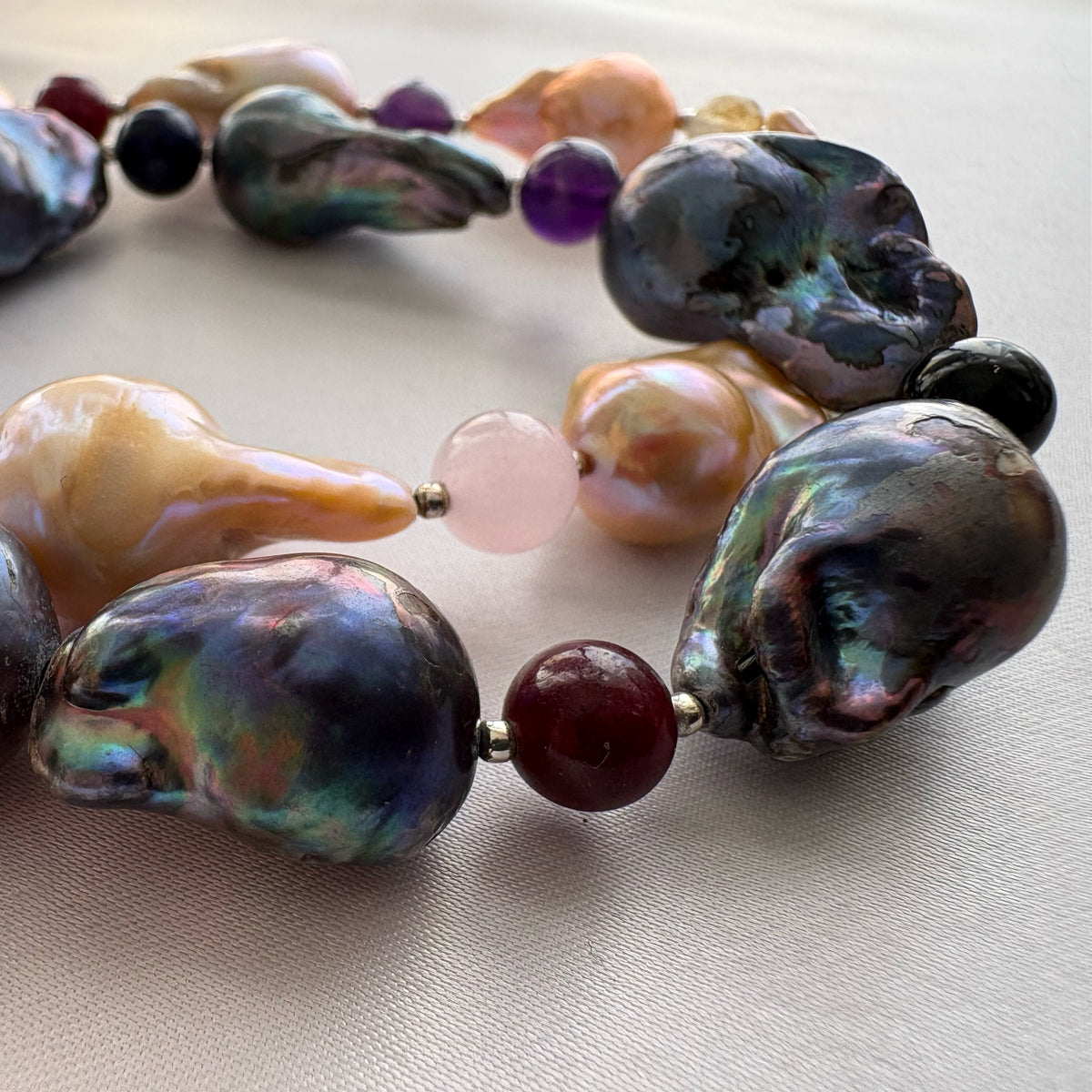 Close-up of a deep red gemstone bead between two large iridescent dark blue-grey keshi pearls, with golden-cream pearls and pale pink and purple beads behind them on cream fabric.