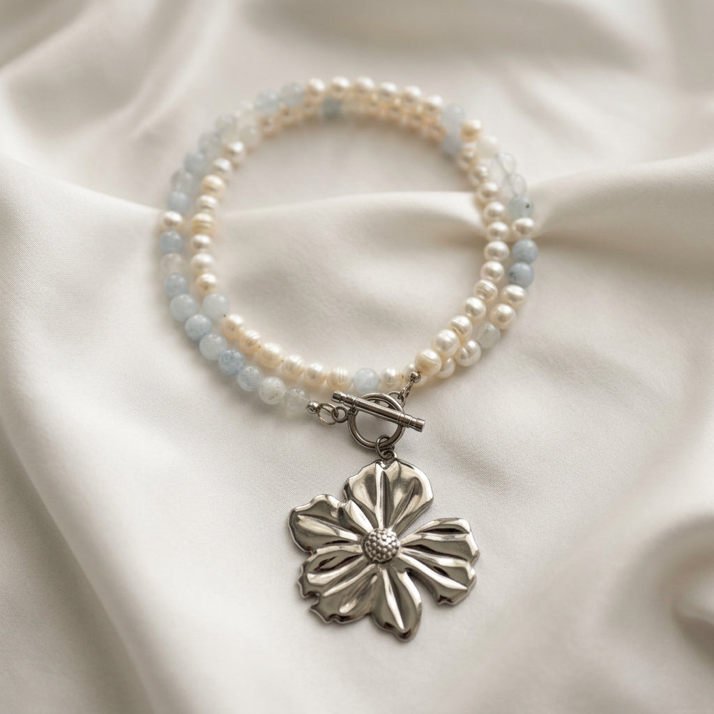 White baroque pearl and pale blue aquamarine necklace arranged in overlapping loops on cream fabric, with a fastened stainless steel toggle clasp and large silver-coloured flower pendant.