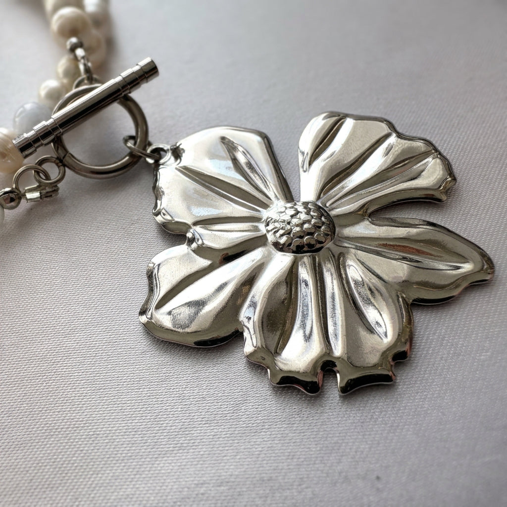 Close-up of a polished silver-coloured five-petal flower pendant with sculpted petals and a textured centre, beside a fastened stainless steel toggle clasp on cream fabric.