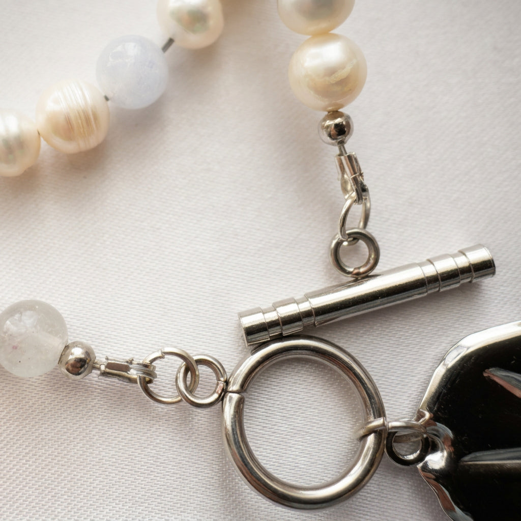 Close-up of an unfastened stainless steel toggle clasp, showing the separate bar and circular ring, connecting rings and bead ends beside white baroque pearls and pale blue aquamarine on cream fabric.