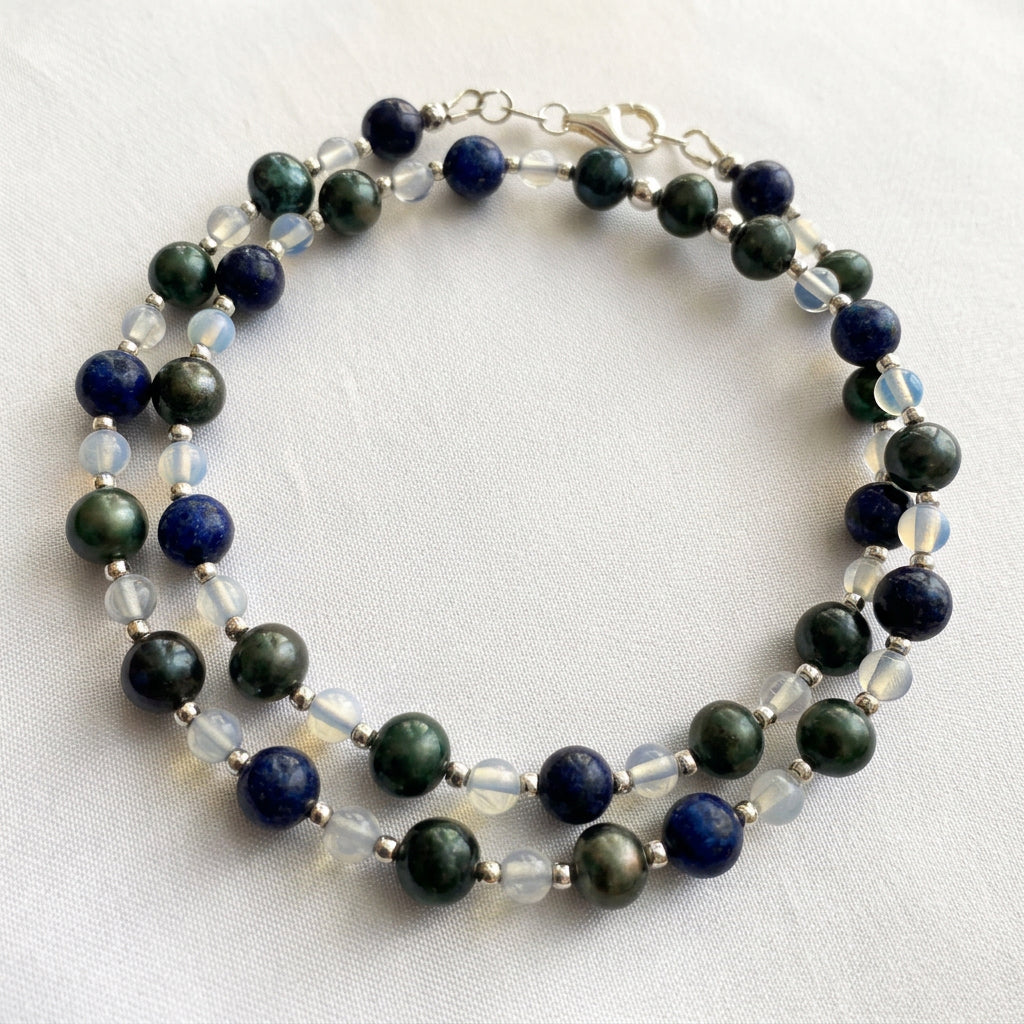 Peacock pearl, lapis lazuli and moonstone necklace loosely coiled on cream satin, showing dark green pearls, deep blue stones, pale translucent beads and small sterling silver spacers, with the clasp near the top.